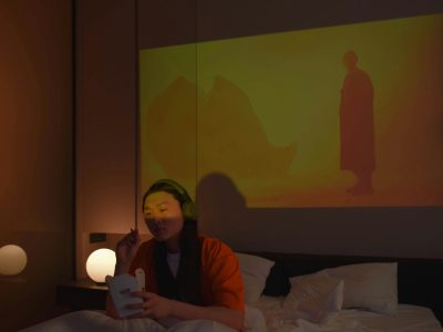 An Asian woman with headphones enjoys eating on bed watching a movie projection.